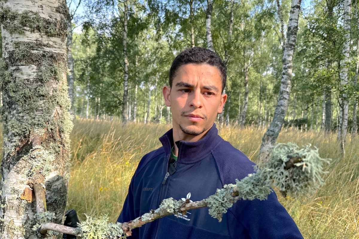 Man i blå jacka står vid björk med lavar på. Foto.